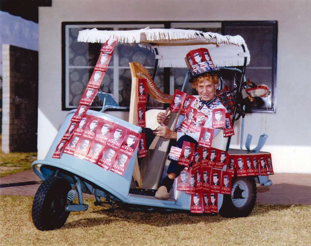 Photo of Harpo Marx in a motor cart decorated with Kennedy campaign posters