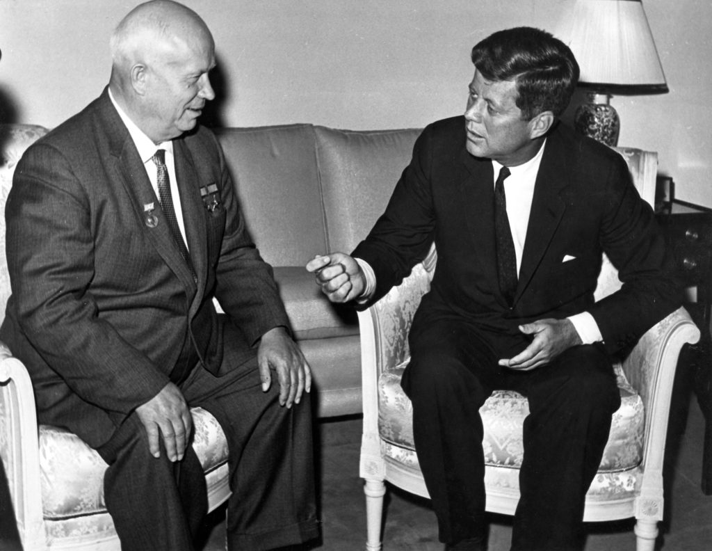 Black-and-white photograph featuring Soviet Premier Nikita Khrushchev, seated at left, looking at President John F. Kennedy, seated at right, as Kennedy gestures while speaking.