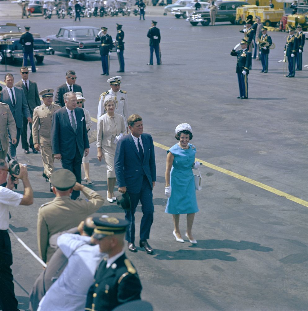 Color photograph of President John F. Kennedy and Rose Fitzgerald Kennedy walking outdoors with a group of people following, and members of the press and military looking on.