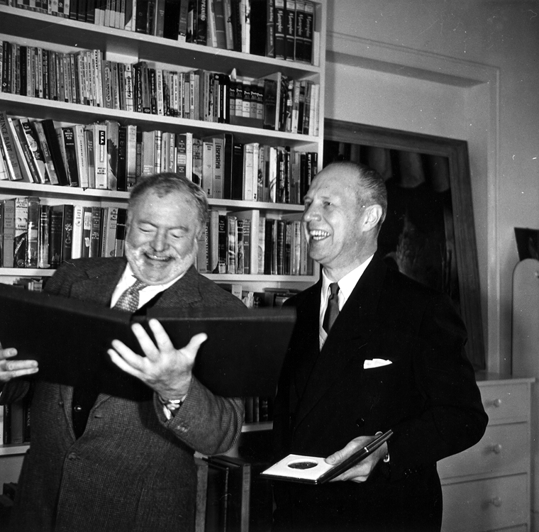 A black-and-white image of the writer Ernest Hemingway smiling while looking at an open binder as he stands next to Sweden's Ambassador to Cuba, Per Gunnar Vilhelm Aurell, who holds a medal in a small case. The two men stand in front of a tall bookshelf filled with books.